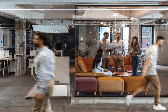 Arbeitende Menschen in einem modernen Büro, drei davon stehen und sprechen, zwei Menschen sind in Bewegung