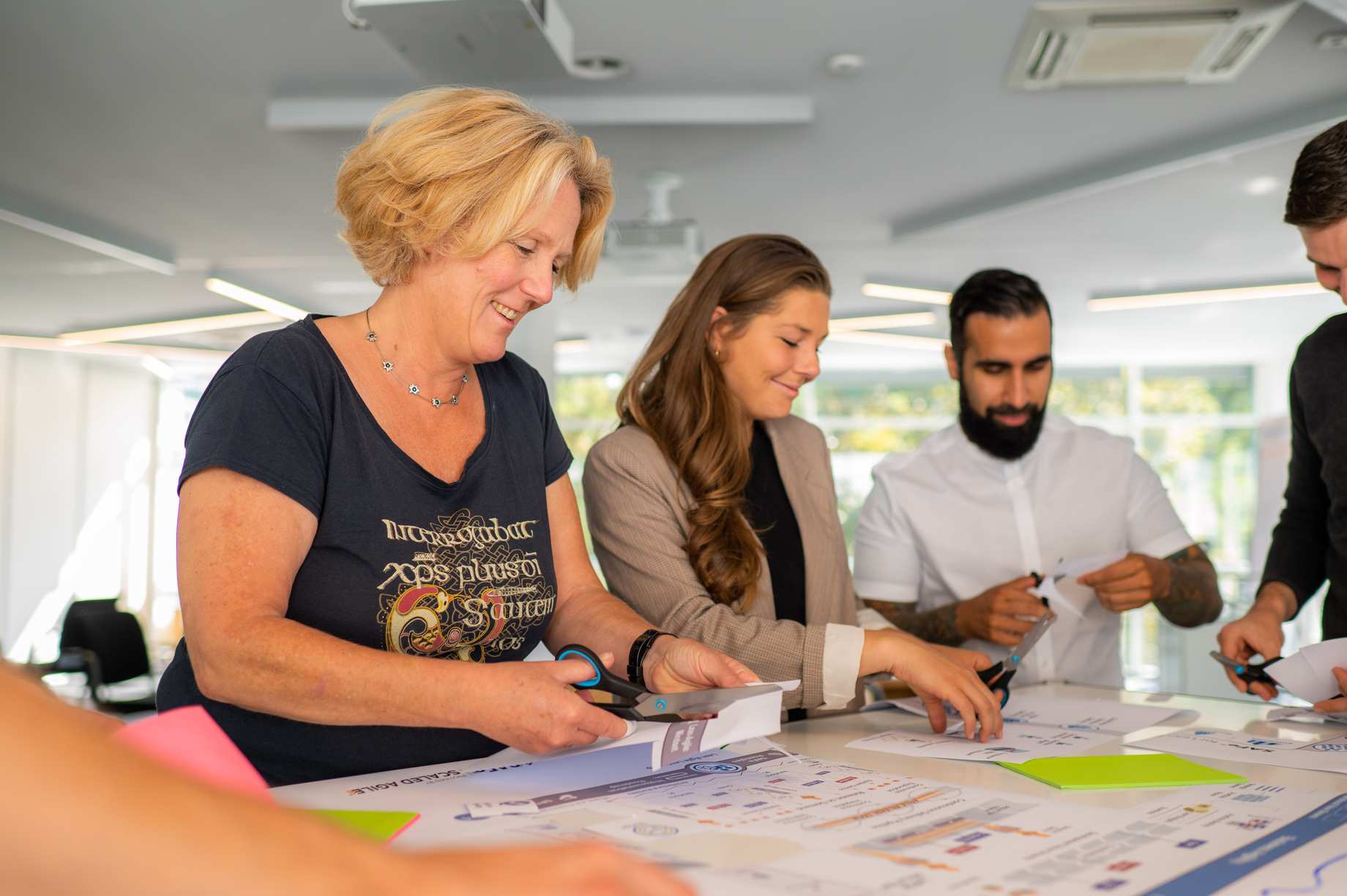 A group of 4 laughing people stand around a table and cut out elements from a SAFe poster.