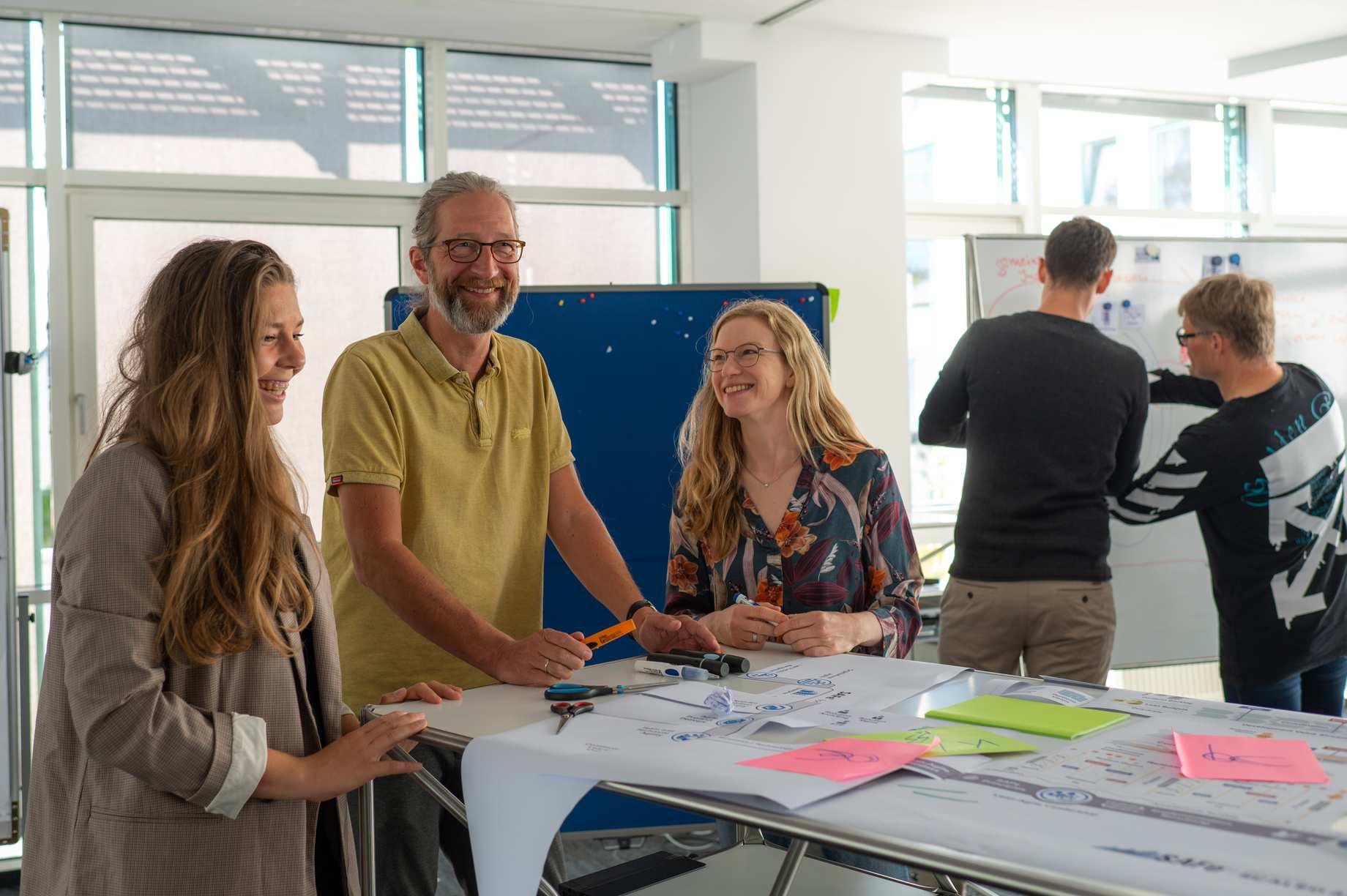 Links im Vordergrund steht eine Gruppe von 3 lachenden Personen um einen Tisch und diskutiert. Rechts im Hintergrund arbeiten 2 Personen miteinander an einem Whiteboard.