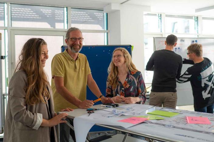 Links im Vordergrund steht eine Gruppe von 3 lachenden Personen um einen Tisch und diskutiert. Rechts im Hintergrund arbeiten 2 Personen miteinander an einem Whiteboard. 