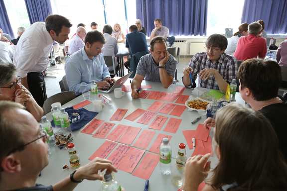 Many people sit in table groups in a large room. Each table group works together and has working material on the table.