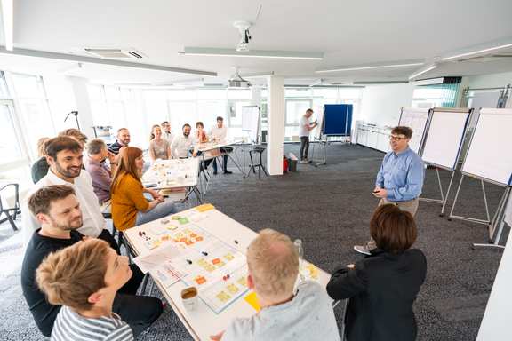 People in a large room working at high tables. A trainer stands in front and explains. A second trainer takes notes on the flipchart.