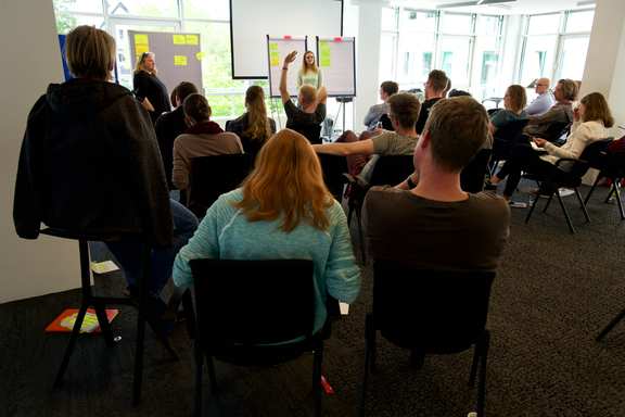 Eine große Gruppe von menschen in einem Saal. Vorne steht eine Person und moderiert.
