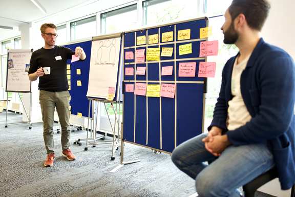 Zwei Personen moderieren ein Meeting. Sie stehen vorne im Raum, neben einer Tafel mit der Agenda.