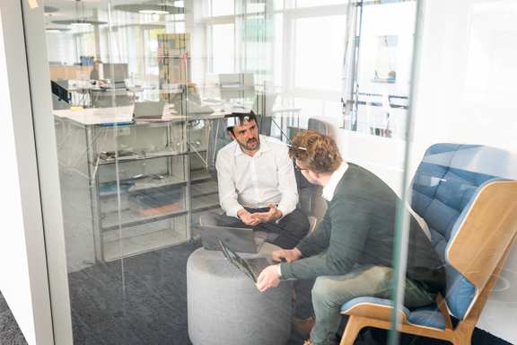 Zwei Personen sitzen in einem Raum und reden miteinander. Sie sind durch eine Glasscheibe sichtbar.
