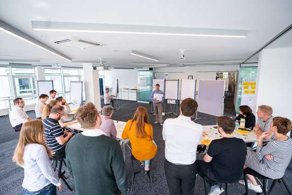 A large room with people at bar tables, a trainer in front; view from back to front to the trainer