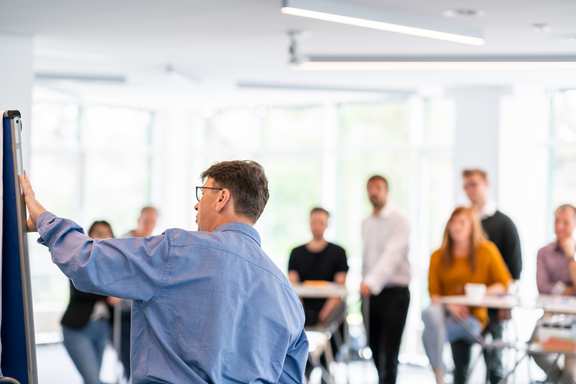 a trainer explains something on the flipchart and many training participants listen spellbound